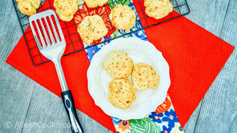 Cheddar Bay Biscuits in a white bowl, sitting on a red table cloth with additional biscuits in the background, sitting on a wire rack