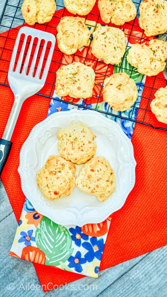 Bird’s eye view of cheddar biscuits in a white bowl, with additional biscuits in the background, sitting on a cooling rack.