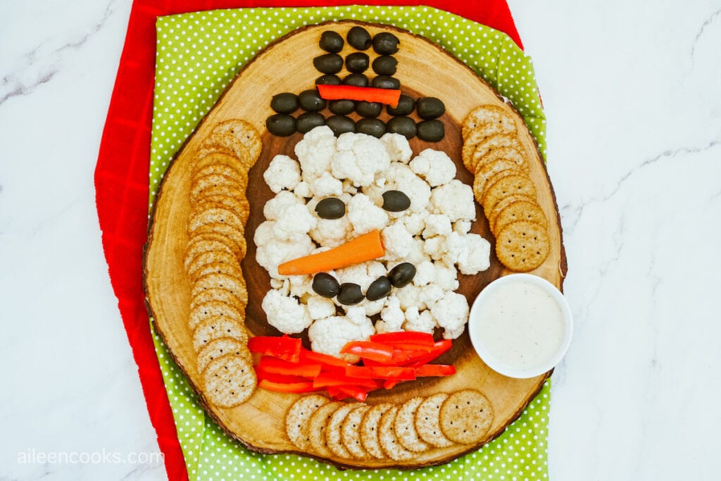 A bird’s eye view of a snowman vegetable platter on a wooden tray, sitting on red and green placemats