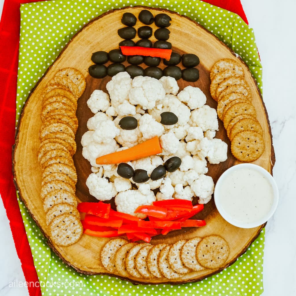 Aerial view of cauliflower in the shape of a snowman, with black olives, a carrot and red pepper. The veggies sit on a wooden platter on a red and green placemat