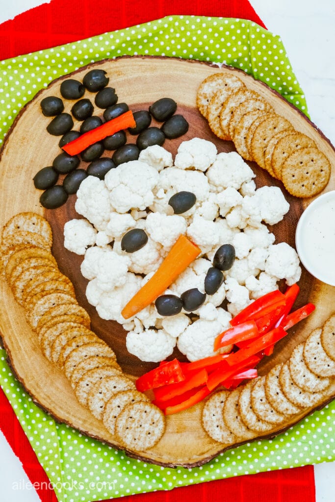 Cauliflower, black olives, and red bell peppers served on a wooden tray to look like a snowman, surrounded by crackers and ranch sauce.
