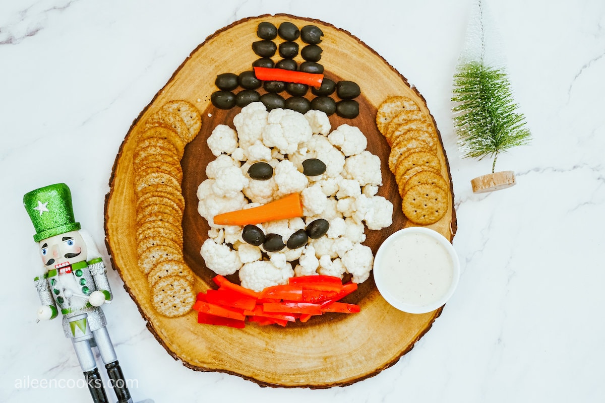 Cauliflower, black olives, and red bell peppers served on a wooden tray to look like a snowman, surrounded by crackers and ranch sauce, sitting on a marble countertop with some holiday décor.