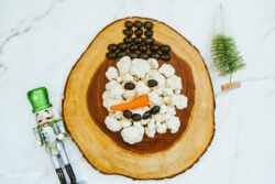 A Snowman Veggie Tray being assembled on a wooden tray, with cauliflower, olives and a carrot being used as the snowman’s face