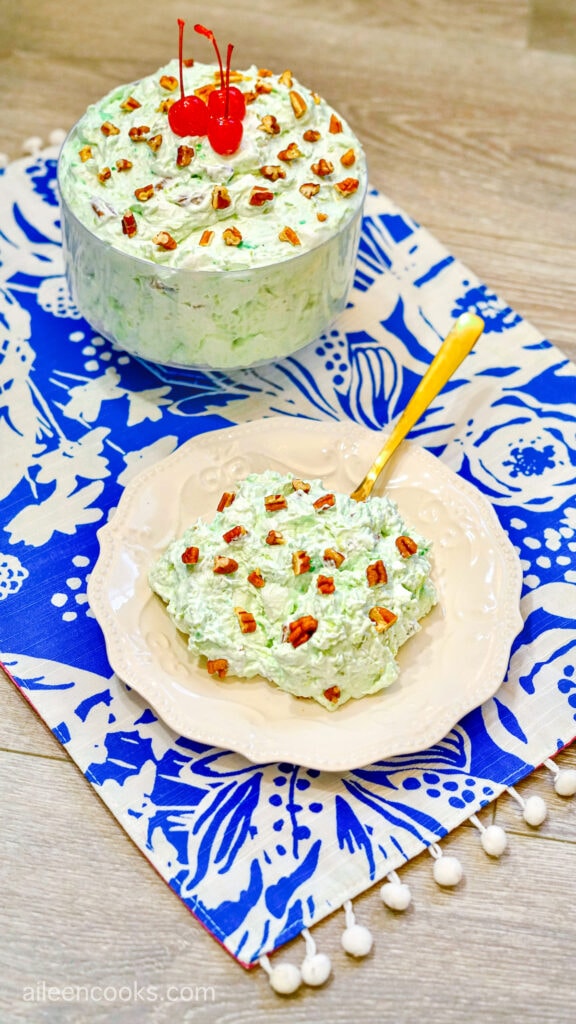 A bowl of Watergate Salad, garnished with chopped almonds, next to a large bowl filled with more, sitting on a patterned tea towel.
