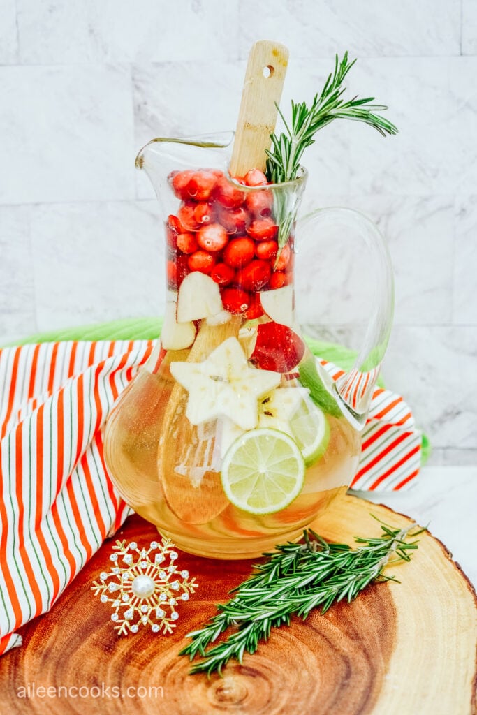 A large glass pitcher filled with champagne sangria, sitting on a wooden cutting board, with a striped tea towel in the background