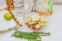 Adding fruits like apples and cranberries into a large pitcher, with sprigs of rosemary on the counter