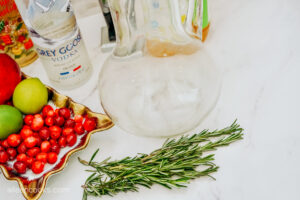 Sprigs of fresh rosemary, a bottle of Vodka, and fruit on a white countertop