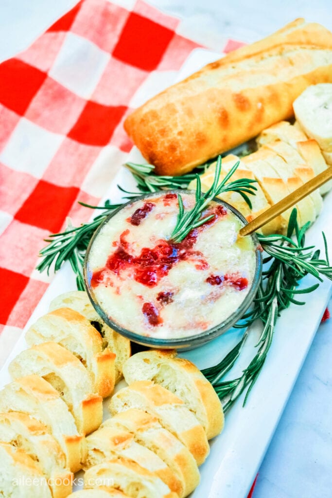 Aerial view of baked brie, mixed with cranberry sauce and fresh rosemary, surrounded by slices of baguette, sitting on a red and white plaid tea towel