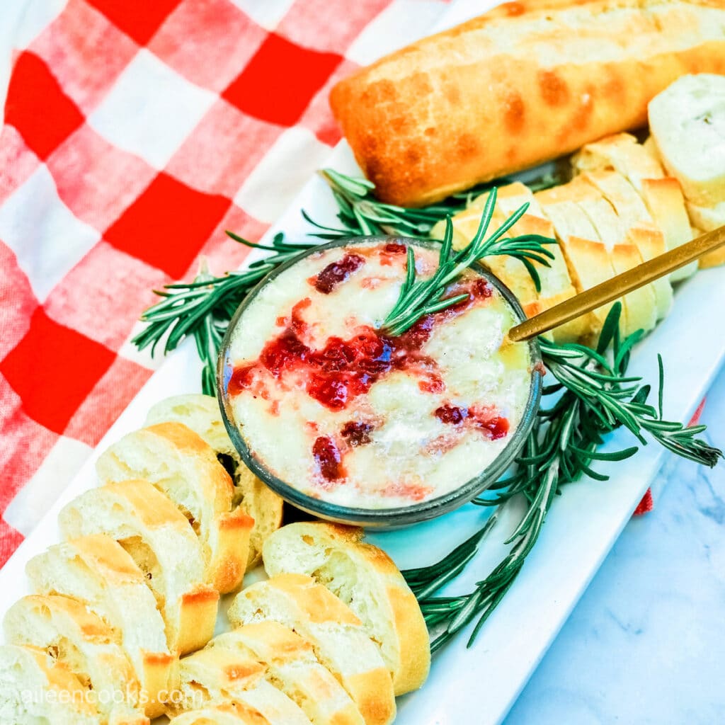 Cranberry baked brie, served on a long rectangular platter, surrounded by slices of baguette, sitting on a red and white plaid tea towel