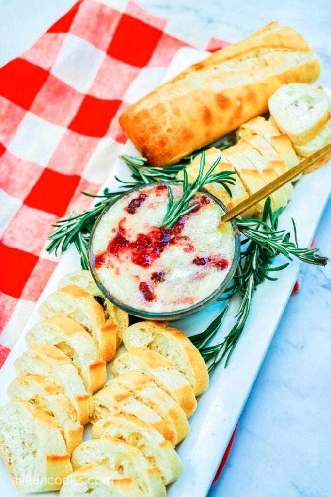 Aerial view of cranberry baked brie, with a sprig of fresh rosemary and served with baguette slices.