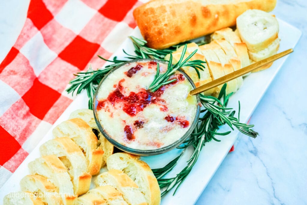 Cranberry baked brie, served on a long rectangular platter, surrounded by slices of baguette, sitting on a red and white plaid tea towel