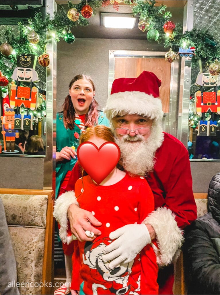 A girl posing with Santa Clause, with a woman dressed as an elf in the background.