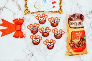 Homemade Reindeer donuts on a glass serving dish, sitting on a marble countertop and surrounded by additional ingredients, including a bag of pretzels