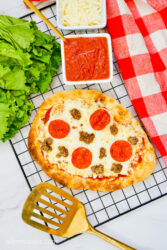 Grilled Flatbread Pizza on a wire rack, surrounded with a gold spatula, red tea towel, and additional pizza ingredients.