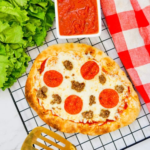 Grilled Flatbread Pizza on a wire rack, surrounded with a gold spatula, red tea towel, and additional pizza ingredients.