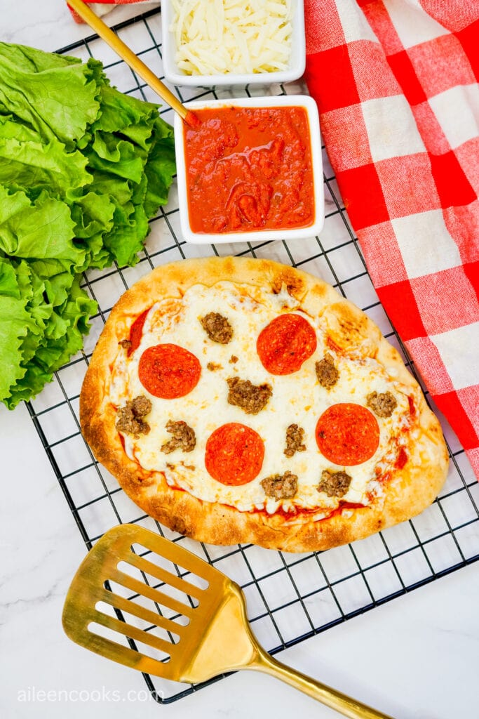 Grilled Flatbread Pizza on a wire rack, surrounded with a gold spatula, red tea towel, and additional pizza ingredients.