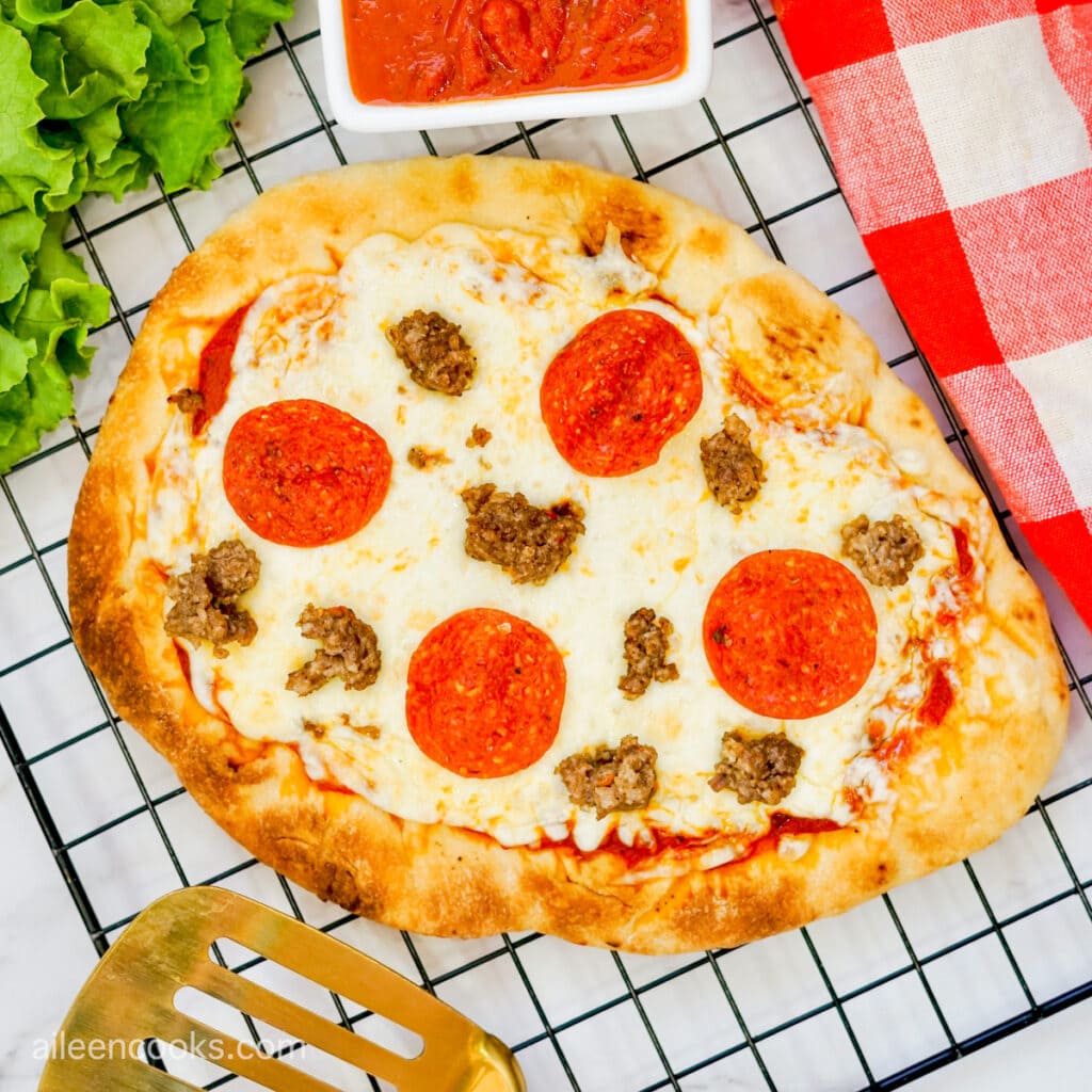 A closeup shot of a flatbread pizza, sitting on a black wire rack, topped with pepperoni and sausage