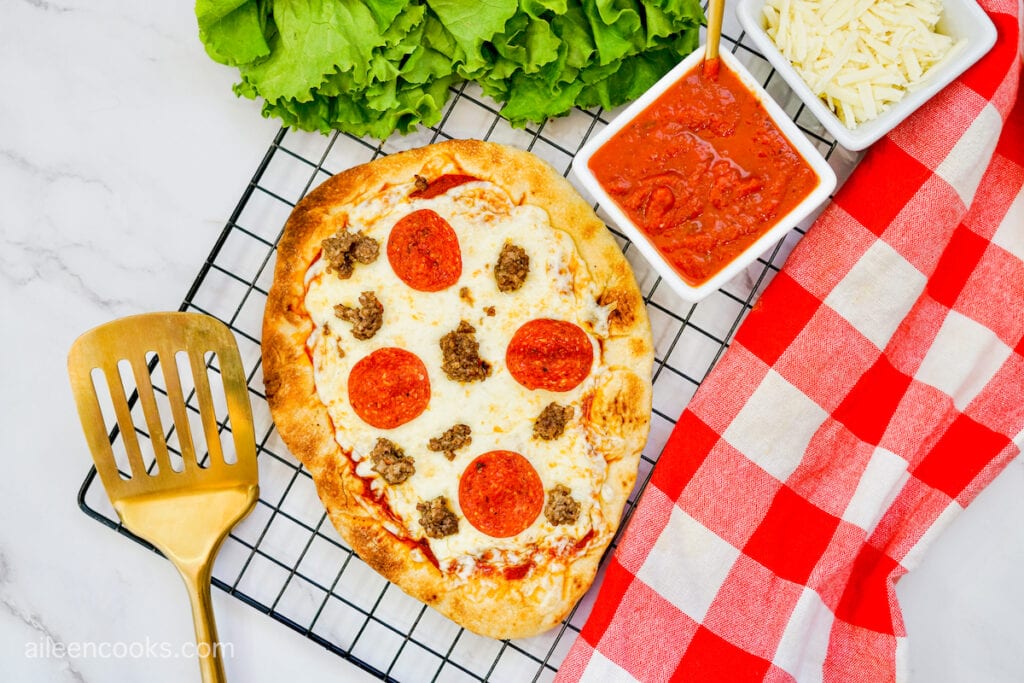 Grilled Flatbread Pizza on a wire rack, surrounded with a gold spatula, red tea towel, and additional pizza ingredients.