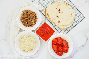 Ingredients laid out on a marble countertop, in white bowls, to make Grilled Flatbread Pizza