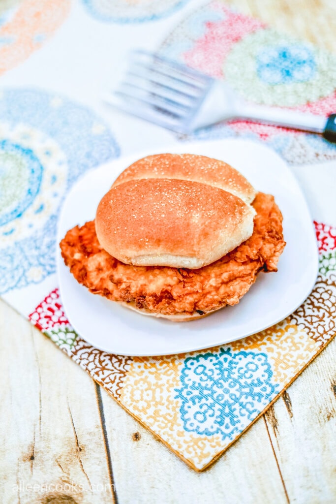 A Crispy Chicken Sandwich, sitting on a white plate, on colorful place mats, ready to be served.