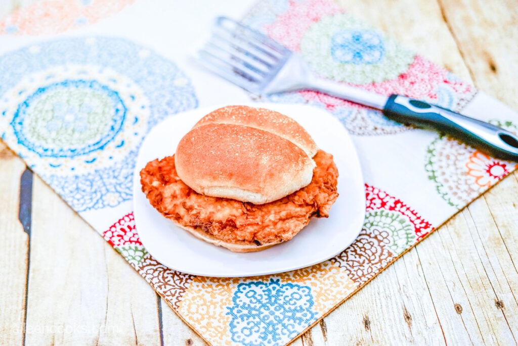 A serving of homemade fried chicken sandwich, on a square, white plate – sitting on a colorful place mat.