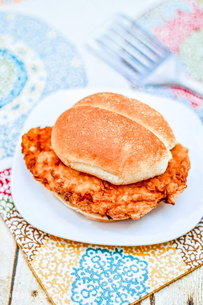 Fried chicken breast, sandwiched between two burger buns and served on a white plate. The plate itself sits on a wooden table with some place mats and a spatula.
