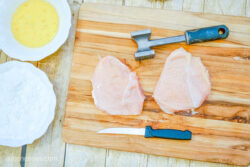 Using a meat mallet to tenderize two pieces of chicken breast on a wooden cutting board
