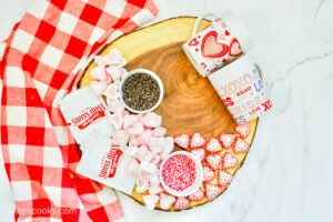 A wood slab topped with bowls of mini chocolate chips and sprinkles, and loose chocolates, heart-shaped marshmallows, and hot cocoa packets.