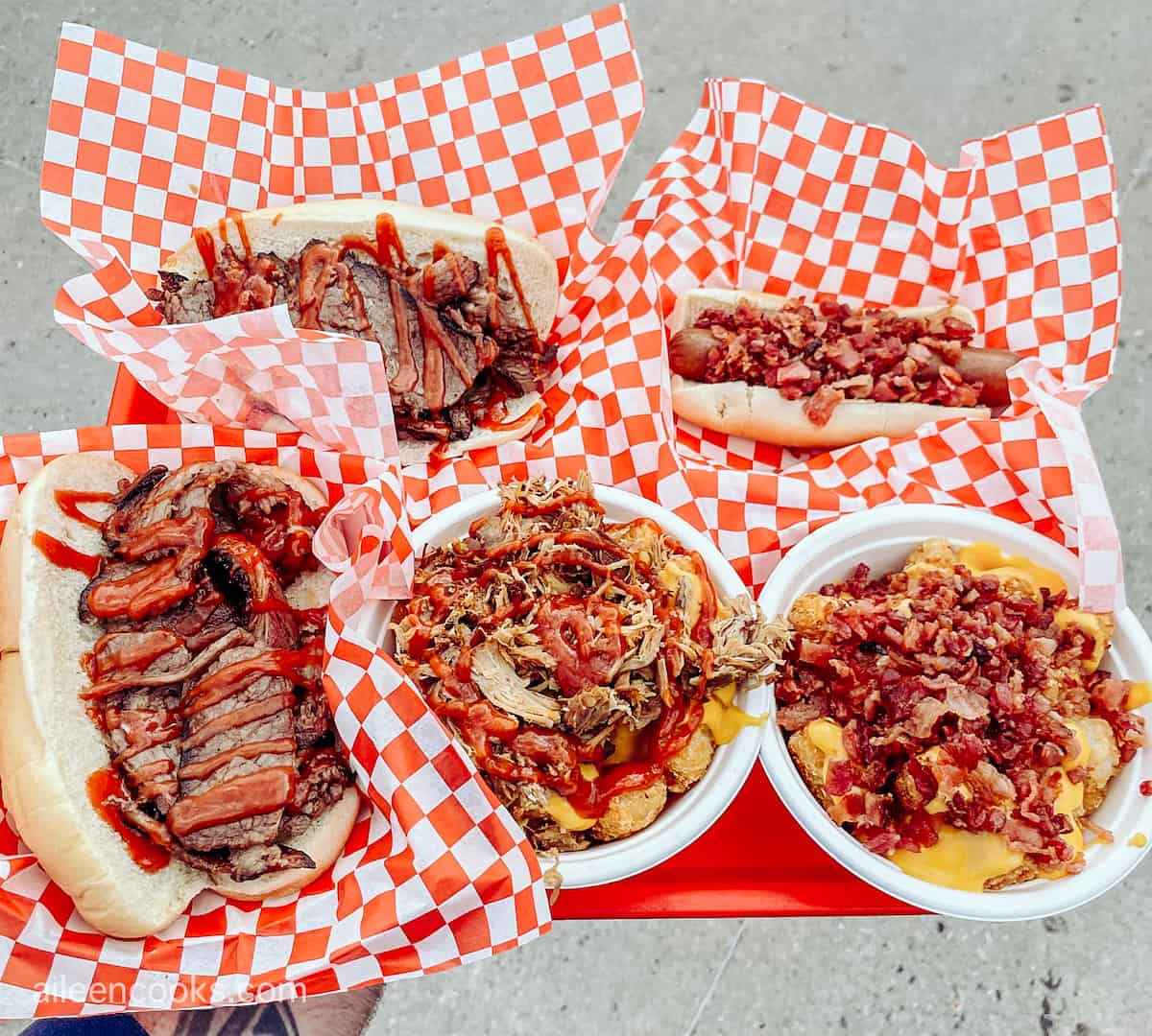 A red tray filled with a variety of barbecue items including pulled pork bowls and bacon topped hot dogs.